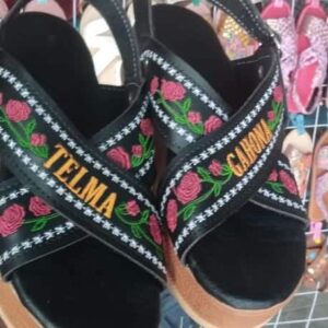 Black sandals with colorful floral embroidery and orange text on the straps (Telma and Calina) shown on display racks.