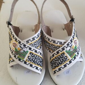 Pair of white open-toe sandals with slingback straps and embroidered black-and-white geometric panels, featuring small religious figures on the sides.